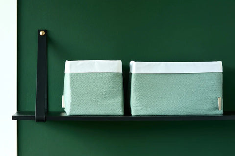 Zwei hellgrüne Stoffbehälter mit weißem Deckel stehen ordentlich auf einem minimalistischen schwarzen Regal vor einer dunkelgrünen Wand.