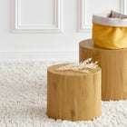 Zwei zylindrische Holzhocker auf einem weißen Teppich; einer hält das KraftKids Körbchen rund Leinen gelb senfgelb, ideal für Aufbewahrung Wickeltisch, während der andere eine kleine getrocknete Pflanze hat. Im Hintergrund ist eine weiß getäfelte Wand zu sehen.