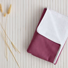 Eine KraftKids Wickelunterlage Leinen magenta dunkel wird mit einer weißen Ecke gefaltet und auf eine weiße Lattenunterlage neben getrocknete beige Grashalme gelegt.