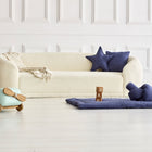 Ein gemütliches weißes Sofa mit einer Decke, marineblaue Stern- und Knotenkissen, eine KraftKids Krabbeldecke Leinen blau dunkelblau Spielmatte mit Bauklötzen, Leinen Körbchen zur Aufbewahrung und ein Spielzeugflugzeug aus Holz in einem hellen, weiß getäfelten Zimmer.