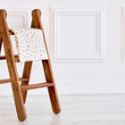 Eine KraftKids Babydecke für Babyschale Sommer Musselin Zweige braun liegt gefaltet über einer Stufe einer kleinen Holzleiter, die auf weißem Boden vor einer weißen Wand steht.
