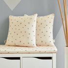 Zwei cremefarbene KraftKids Kissenbezug Musselin Kirschen mit Kirschenmuster liegen auf einer passenden Bank mit Aufbewahrungsschubladen. Eine hohe Vase mit Bambusstäben steht in der Nähe vor einer grauen, geometrischen Wand.