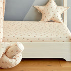 Ein gemütliches Kinderbett mit dem KraftKids Spannbettlaken Musselin Kirschen, einem pfirsichfarbenen Spannbetttuch mit zartem Kirschdruck und einem sternförmigen Kissen, neben einem hellen Holzboden und einer zartgrauen Wand.