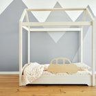 Das KraftKids Rausfallschutz aus Leimholz Auto ist ein minimalistisches Kinderbett in Form eines Hausrahmens mit einem ausgeschnittenen Auto aus Holz, weißem und rosa Bettzeug, auf einem Holzboden vor einem grauen Wandbild.