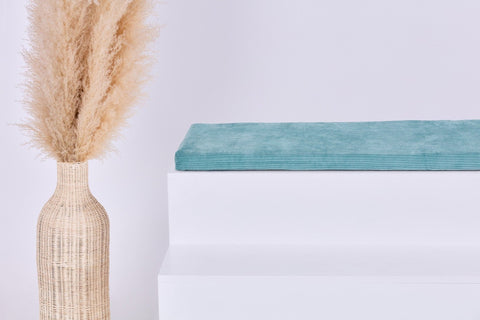 Eine Weidenvase mit beigem Pampasgras steht neben einem weißen Stufenpodest, auf dem ein gefaltetes tealfarbenes Handtuch und die KraftKids GmbH Sitzauflage für Ikea STUVA/SMÅSTAD Cord Breitcord Mint vor einem schlichten hellen Hintergrund liegen.