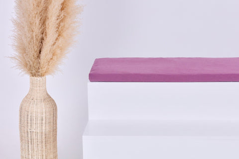Eine hohe geflochtene Vase mit beigem Pampasgras steht neben einer modernen weißen Bank mit der KraftKids GmbH Sitzauflage für Ikea STUVA/SMÅSTAD Musselin purpur vor einem minimalistischen hellen Hintergrund.