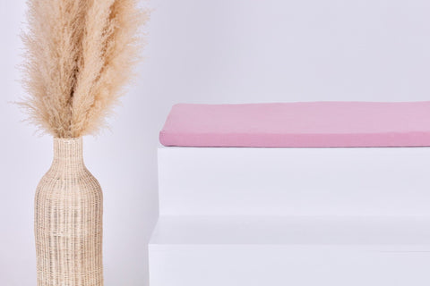 Eine hohe geflochtene Vase mit getrocknetem Pampasgras steht neben einem weißen Stufenpodest, auf dem die KraftKids GmbH Sitzauflage für Ikea KALLAX 2er, 3er und 4er Regal in Musselin rosa vor einem schlichten hellen Hintergrund steht.