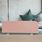 Ein minimalistisches KraftKids-Bett mit einem hellen Holzrahmen ist mit dem Bettschutzgitter Rausallschutz in Pastellrosa, grünen Sternkissen und einem weißen Kissen ausgestattet und steht vor einer kahlen Betonwand.