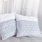 Zwei KraftKids Kissenbezug "weiße Pfeile auf Grau" liegen auf einem weißen Metallbettgestell auf einer hellgrauen Strickdecke, mit weißen Vorhängen im Hintergrund.