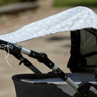 Eine Nahaufnahme eines Kinderwagens, der mit dem KraftKids Sonnensegel weiße Pfeile auf Grau ausgestattet ist, zeigt das weiße Pfeilmuster auf hellgrauem Stoff, das sich über den Sitz im Freien erstreckt, mit einem unscharfen Steinweg im Hintergrund.