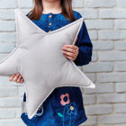 Ein kleines Kind in einem blauen Kleid hält ein KraftKids Sternkissen Unigrau vor einer weißen Backsteinmauer, was es zu einer charmanten Ergänzung für jedes Kinderzimmer macht. Das Gesicht des Kindes ist nicht sichtbar.