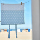 Der KraftKids Bezug für Wickeltischauflage weiße Diamante auf Pastell Blau (75 x 70 cm) hängt an einer Wäscheleine mit Bändern, vor einem sonnigen Strand mit Hütten und blauem Himmel - ideal als stilvoller Wickelauflagenbezug.
