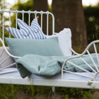 Ein weißes Metallbett mit KraftKids Bettwäscheset Musselin mint aus 100% Baumwolle und Kissen, darunter ein lächelndes, kronenförmiges Kissen, steht auf einer Wiese im Freien mit Bäumen und Grünzeug für erholsamen Schlaf.