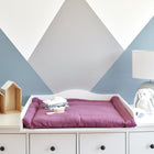 Eine weiße Kommode mit einer KraftKids Wickelauflage Musselin purpur enthält Babykleidung und Toilettenartikel. Auf der linken Seite stehen kleine hausförmige Regale mit Plüschtieren, rechts eine Lampe und ein Plüschhase. Die Wand zeigt ein blau-weißes geometrisches Muster.