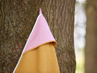 Ein KraftKids Kapuzenhandtuch "weiße Punkte auf Koralrosa Waffel Piqué senf" hängt an seiner Schlaufe an einem Baumstamm und zeigt seinen weichen senfgelben Waffelstoff und die rosa Kapuze mit weißen Punkten.