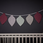 Die Wimpelkette Musselin purpur grau mint Pusteblumen von KraftKids schmückt als Deko das Kinderzimmer und hängt über einem weißen Babybett vor einer dunklen Wand.