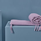 Eine gefaltete und teilweise gerollte KraftKids Bettumrandung Nestchen Waffel Piqué rosa aus 100% Baumwolle mit Bindebändern liegt auf einem hellblauen rechteckigen Sockel vor passendem Hintergrund.