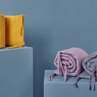 Drei gerollte KraftKids Bettumrandung Nestchen Waffel Piqué grau mit Krawatten sind auf blauen Sockeln vor blauem Hintergrund ausgestellt - ein gelber Artikel auf der linken Seite und lavendelfarbene und graue Artikel auf der rechten Seite.