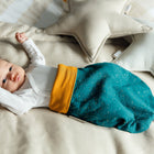 Ein Baby liegt auf einem beigen Bett mit sternförmigen Kissen, trägt ein weißes Oberteil und den KraftKids Strampelsack Herbst Winter Musselin goldene Punkte auf Petrol, kuschelig und gemütlich in dem weichen Schlafsack.