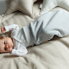 Ein Baby, bekleidet mit einem weißen langärmeligen Strampelsack und dem KraftKids Strampelsack Herbst Winter Musselin grau Pusteblumen, liegt auf einem beigen Bett mit beigen und sternförmigen Kissen im Hintergrund.