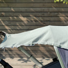 Ein Sonnensegel Musselin mint von KraftKids ist über den Griff und das Verdeck eines Kinderwagens drapiert, der im Freien vor einem Holzzaun steht, während das Sonnenlicht Schatten wirft.