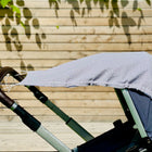 Ein KraftKids Sonnensegel Musselin grau Punkte spendet Schatten für das Baby auf einem Kinderwagen mit braunem Griff. Im Freien erzeugt das Sonnenlicht Blätterschatten auf einem Holzzaun - ein idealer Sonnenschutz fürs Baby.