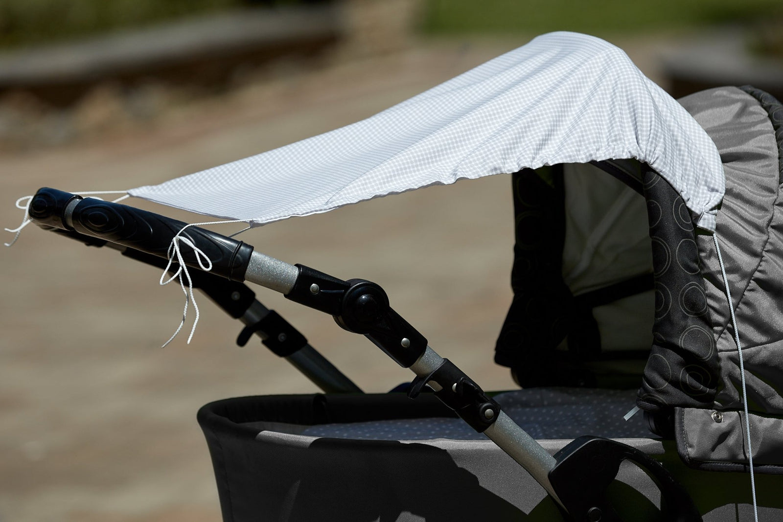 Eine Nahaufnahme zeigt einen Kinderwagen, über dessen Verdeck und Griffe das KraftKids Sonnensegel Karo grau drapiert ist, das den Kinderwagen effektiv vor der Sonne schützt. Der sanft verschwommene Hintergrund hebt den beschatteten Kinderwagen hervor.