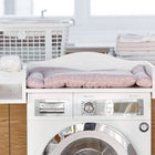 Ein KraftKids Wickelaufsatz für Waschmaschinen und Trockner mit pinker Auflage steht auf der Waschmaschine. Daneben: Ablageregal mit Handtüchern, einem Wäschekorb und Feuchttüchern im modernen, hellen Waschraum.