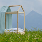 A KraftKids Bettbaldachin Musselin mit goldenen Punkten auf Grün ziert ein minimalistisches Holzhausbett inmitten von Wildblumen, vor Bergen und bewölktem Himmel - ein traumhafter Hingucker fürs Kinderzimmer.