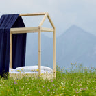 Ein KraftKids Bettbaldachin Musselin dunkelblau und weißes Bettzeug schmücken ein Holzhausbett in einer Wildblumenwiese, Berge und Wolken im Hintergrund - die perfekte Inspiration für Kinderzimmer Deko.