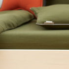 Eine Nahaufnahme eines grünen Kissens und eines Kissens auf einem hellen Holzsofa, mit einem orangefarbenen Kissen im Hintergrund. Die gemütliche Szene ist mit dem KraftKids Spannbettlaken Doppelkrepp Grün Herbstgrün gestaltet, das die satte Farbe und Textur des Stoffes hervorhebt.