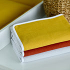 Drei ordentlich gefaltete Handtücher in Gelb, Rost und Weiß mit weißem Rand sitzen neben einem Korb und einem Kissen - eine ideale Ergänzung zur KraftKids Wickelunterlage Doppelkrepp Gelb Senf, um Ihr Kinderzimmer stilvoll zu organisieren.