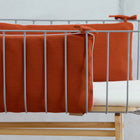 Eine Nahaufnahme eines modernen Babybettes mit hellgrauen Metallstäben, einem KraftKids Bettumrandung Nestchen Doppelkrepp Rot Herbstrot an den Seiten und einer weißen Matratze auf einem Holzrahmen vor einer hellgrauen Wand.