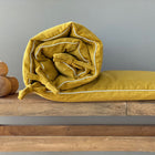 Die Bettumrandung Nestchen Cord Breitcord Gelb Mustard von KraftKids, ein handgefertigter senfgelber Kinderbett-Stoßfänger, wird auf einer Holzbank neben drei runden Holzobjekten vor einer schlichten grauen Wand aufgerollt und verschnürt.