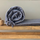 Die KraftKids Bettumrandung Nestchen Cord Breitcord Grau aus 100% Baumwolle mit weißer Einfassung und Stoffbändern liegt aufgerollt auf einem Holztisch neben drei runden Holzkugeln und einem passenden grauen Kissen vor einem neutralen Hintergrund.