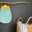 Ein kleiner mintfarbener Cord KraftKids Strampelsack Herbst Winter Cord Breitcord hängt an einem hölzernen Ast an einer dunklen Wand. Daneben steht ein geflochtener Korb mit gerollten Decken und Handtüchern.