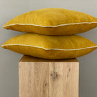 Zwei KraftKids Kissenbezüge Cord Breitcord Gelb Mustard mit weißer Paspelierung sind auf hellem Holz vor einem grauen Hintergrund gestapelt, perfekt, um Ihrer Bettwäschesammlung einen Farbtupfer zu verleihen.