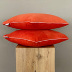 Zwei KraftKids Kissenbezüge Cord Breitcord Orange Reinorange mit weißer Paspel sind auf einem Holzhocker gestapelt und stehen vor einem einfarbig beigen Hintergrund.