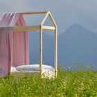 Ein kleines Himmelbett aus Holz mit weißem Bettzeug und einem rosa Bettbaldachin Musselin goldene Punkte auf Rosa von KraftKids steht in einem Feld mit Wildblumen, mit dunstigen Bergen und einem wolkenverhangenen Himmel im Hintergrund.