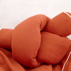 Ein plüschiger, rostfarbener KraftKids Wickeltischumrandung Doppelkrepp Rot Herbstrot ruht auf passender Bettwäsche mit weißer Paspel, ergänzt durch eine weiche Bettrolle, vor einer neutralen hellen Wand.