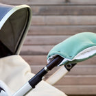 Eine Nahaufnahme des KraftKids Kinderwagenmuff Doppelkrepp Grün Jade, handgenäht und wärmeisoliert, am Kinderwagenlenker vor einem Holzzaun. Der Sitz ist teils zu sehen und mit einer weißen Decke bedeckt.