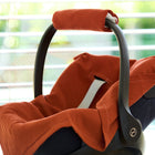 Eine KraftKids Babydecke für Babyschale Sommer Doppelkrepp Rot Herbstrot bedeckt einen schwarzen Kinderautositz, der im Innenbereich an einem Fenster mit horizontalen Jalousien steht und in natürliches Licht getaucht ist.