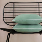 Ein schwarzer Metallstuhl mit gerasterter Rückenlehne hält zwei gestapelte KraftKids Kissenbezug Doppelkrepp Grün Jade mit weißer Paspel vor einer beigen Wand.