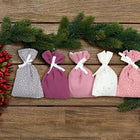 Fünf kleine KraftKids Adventskalender Musselin Rosa Purpur Weiß Grau Beutel mit weißen Bändern sind auf Holz arrangiert, umgeben von Tannenzweigen und roten Beeren - ideal für Ihre festliche Dekoration.