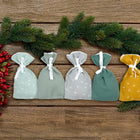 Vier kleine KraftKids Adventskalender Musselin Gelb Grau Taschen in Pastellgrün, Grau und Gelb sitzen auf einer Holzfläche mit Tannenzweigen und roten Beeren für ein festliches Weihnachtsarrangement.