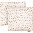 Zwei quadratische KraftKids Kissenbezüge Musselin Zweige braun aus unserer Bettwäschekollektion mit weißem Hintergrund, kleinen braunen Blattmustern und einem Markenetikett werden übereinander gelegt und sorgen so für einen gemütlichen Look.